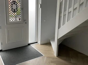 Hal met voordeur, visgraatvloer, schoonloopmat en een witte trap