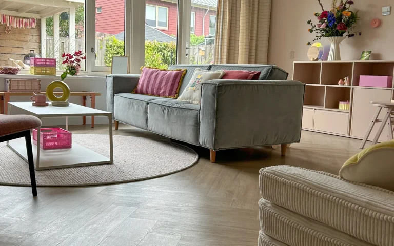 Woonkamer met een grijze bank en een lichte visgraatvloer, met uitzicht op straat