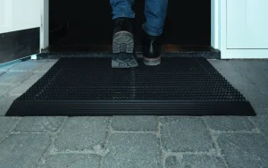 Iemand stapt met schoenen op een zwarte schoonloopmat die in een deuropening op een natuurstenen vloer ligt