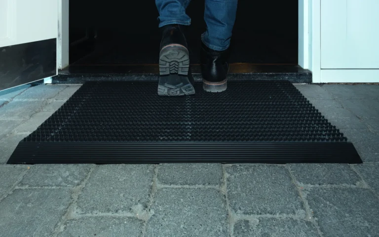 Iemand stapt met schoenen op een zwarte schoonloopmat die in een deuropening op een natuurstenen vloer ligt
